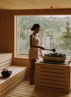 junge Frau gießt Wasser auf heißen Stein in der Sauna foto