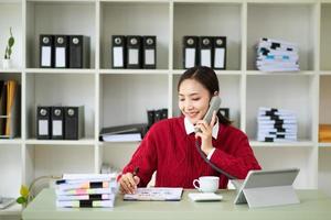 Glückliche asiatische Geschäftsfrau hat die Freude, im Büro auf dem Smartphone, Tablet und Laptop zu sprechen foto