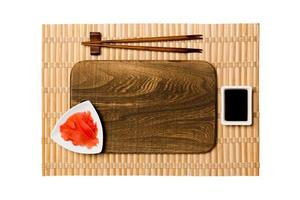 leere rechteckige braune Holzplatte mit Essstäbchen für Sushi, Ingwer und Sojasauce auf gelbem Bambusmattenhintergrund. Draufsicht mit Kopierbereich für Ihr Design foto