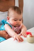 Kind sechs Monate alt mit Mutter in einer Küche an einem Tisch mit Glas mit Gemüsepüree und Löffel. Babymädchen versucht, sich selbst zu essen. foto