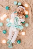 entzückendes lächelndes kleines mädchen in blau-weißem kleid spielt mit weihnachtslichtern auf einem beigen plüschplaid, flach gelegt. foto