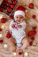 Schönes lächelndes kleines Mädchen im roten Hut des Weihnachtsmanns spielt mit Holzspielzeug auf einem beigefarbenen Plaid mit roten und weißen Weihnachtsdekorationen und Weihnachtslichtern, Draufsicht.