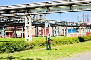 der arbeiter in schutzanzug und maske verwendet ein spezielles gerät, um an einem sonnigen sommertag das gras zu mähen. Rasenmäher in den Händen eines Mannes. vor dem Hintergrund von Rohren zur Chemieproduktion foto