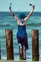 glückliche Performance-Künstlerin in blauem Kleid, beschmiert mit blauen Gouachefarben, tanzt am Strand foto