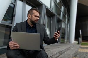 ein mann arbeitet mit einem laptop online und sieht ein handy an, während er in der nähe des bürogebäudes sitzt, online-banking-konzept foto