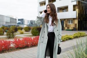 junge, stilvolle Frau in einem Mantel mit Hut, die auf einem Mobiltelefon auf dem Hintergrund von Stadtgebäuden spricht foto