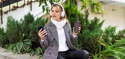 eine erwachsene frau mit kopfhörern wählt einen titel auf einem handy mit einer tasse kaffee in den händen foto