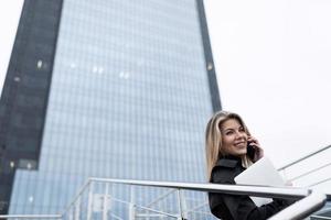 geschäftsfrau blond vor dem hintergrund eines bürohochhauses abgelaufen mit handy und laptop foto