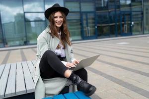 Junge stilvolle Frau, die auf einer Bank mit einem Laptop in einem Hut sitzt und schelmisch in die Kamera lächelt foto