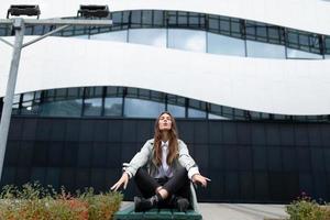 eine junge frau sitzt in einer yogaposition vor der hintergrundkulisse eines bürogebäudes auf einer bank mit geschlossenen augen, die arme ausgebreitet foto