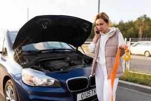 Eine Frau neben einem kaputten Auto telefoniert und hält ein Kabel mit einem Haken zum Abschleppen eines Autos foto