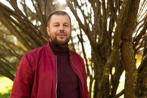 lächelnder junger Mann mit Bart in roter Jacke im Herbstpark foto