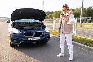 eine fahrerin steht neben einem kaputten auto und ruft technische hilfe auf einem handy an, verkehrsunfallkonzept foto