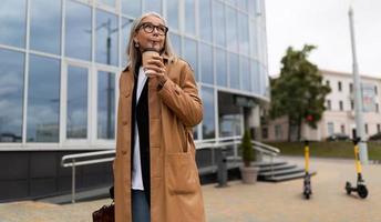 Seniorin geht mit einer Tasse Kaffee in der Hand durch die Stadt und schaut sich vor dem Hintergrund der Glasfassade des Einkaufszentrums um foto