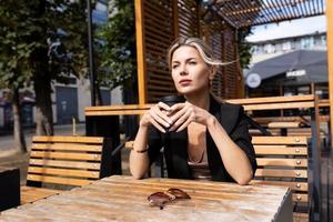 Stilvolle, nachdenkliche Frau mittleren Alters, die Kaffee in einem Café auf der Straße trinkt foto