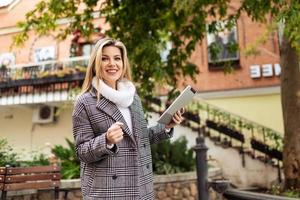 Stilvolle Geschäftsfrau im Herbstmantel mit einem Tablet in den Händen, die mit einem Lächeln auf die Kamera außerhalb des Gebäudes blickt foto