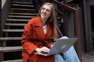 junge Frau, die online an einem Laptop arbeitet, sitzt auf einer Treppe neben einem Café und schaut lächelnd weg foto