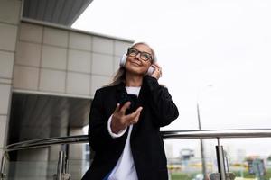 Stilvolle ältere Frau, die mit einem Handy in den Händen Musik in Over-Ear-Kopfhörern hört foto