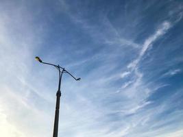 Laternenpfahl mit einer weißen Glühbirne vor einem klaren Himmel. Laterne zur Beleuchtung von zwei Straßen. graue Säule mit Laterne, warmes Gelb. neben der Spur eines fliegenden Flugzeugs foto