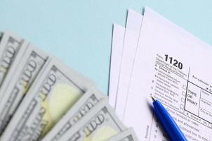 1120 Steuerformular liegt in der Nähe von Hundert-Dollar-Scheinen und blauem Stift auf hellblauem Hintergrund. US-Körperschaftsteuererklärung foto