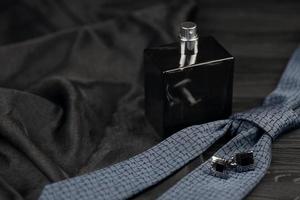 eine flasche mens cologne und manschettenknöpfe mit blauer krawatte liegen auf einem schwarzen luxusstoffhintergrund auf einem holztisch. Accessoires für Herren foto