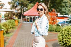 Schöne stilvolle modische Frau in Jeansjacke und Sonnenbrille in einem Vergnügungspark foto