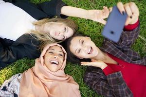 Gemischtrassige Freundinnen lachen, während sie auf dem Rasen liegen und Selfie machen foto