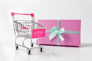 kleiner Supermarkt Lebensmitteleinkaufswagen zum Einkaufen von Spielzeug mit rosa Geschenkbox isoliert auf weißem Hintergrund. verkauf kaufen einkaufszentrum markt shop verbraucherkonzept. Platz kopieren. foto