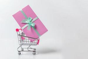 kleiner Supermarkt Lebensmitteleinkaufswagen zum Einkaufen von Spielzeug mit rosa Geschenkbox isoliert auf weißem Hintergrund. verkauf kaufen einkaufszentrum markt shop verbraucherkonzept. Platz kopieren. foto