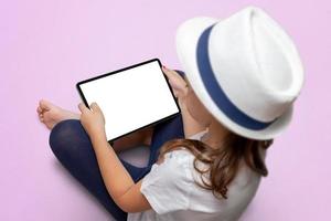 Kleines Mädchen mit weißem Hut sitzt auf einem rosa Boden und hält ein Tablet mit isoliertem Bildschirm. Tablet-Modell foto