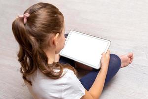Kleines Mädchen, das auf dem Boden sitzt und ein weißes Tablet mit isoliertem Bildschirm hält, um Videospiele, Websites oder Apps zu bewerben foto