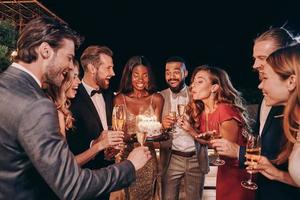 Schöne junge Frau im Abendkleid, die Kerzen auf Kuchen bläst, während sie mit Freunden Geburtstag feiert foto