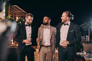 Drei gutaussehende Männer in Anzügen genießen Whiskey und kommunizieren, während sie Zeit auf der Party verbringen foto