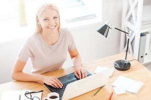 Freude an ihrer Arbeit. Draufsicht einer schönen jungen Frau, die in die Kamera schaut und lächelt, während sie an ihrem Arbeitsplatz im Büro sitzt foto