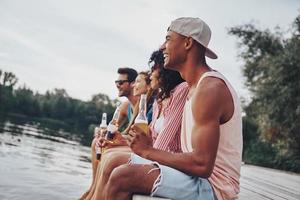 sich frei und entspannt fühlen. gruppe glücklicher junger leute in freizeitkleidung, die lächeln und bier trinken, während sie auf dem pier sitzen foto