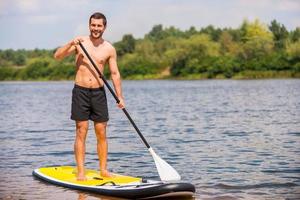 toller tag zum paddeln. hübscher junger Mann, der auf seinem Paddleboard surft und lächelt foto