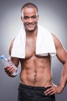 Erfrischung nach dem Training. junger hemdloser afrikanischer mann, der flasche mit wasser hält und lächelt, während er vor grauem hintergrund steht foto