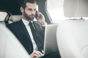 immer verfügbar. hübscher junger Geschäftsmann, der an seinem Laptop arbeitet und telefoniert, während er im Auto sitzt foto