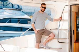 bereit für den Urlaub. lächelnder junger Mann, der den Handlauf hält und in die Kamera schaut, während er auf dem Brett der Yacht steht foto