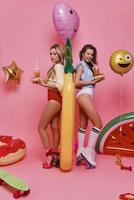 ungesunde Ernährung. in voller Länge von zwei attraktiven jungen Frauen in der Badebekleidung, die Krapfen und Getränke beim Stehen gegen rosa Hintergrund hält foto