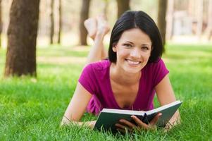 schöner Bücherwurm in der Natur. schöne junge Frau, die Buch liest und lächelt, während sie im Gras liegt foto