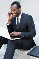 erfolgreicher Geschäftsmann. glücklicher junger afrikanischer mann in formeller kleidung, der am handy spricht und am laptop arbeitet, während er auf der treppe im freien sitzt foto