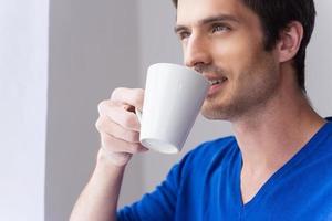 genießt seinen Morgenkaffee. Fröhlicher junger Mann im blauen Pullover, der Kaffee trinkt und wegschaut, während er vor grauem Hintergrund steht foto