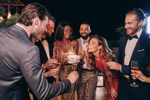 Schöne junge Frau im Abendkleid, die Kerzen auf Kuchen bläst, während sie mit Freunden Geburtstag feiert foto