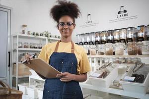 Porträt einer jungen schwarzen Ladenbesitzerin, die lächelt und in die Kamera schaut, die Naturprodukte im Nachfüllladen, Lebensmittel ohne Abfall und plastikfrei, umweltfreundlich mit wiederverwendbaren Behältern arrangiert. foto