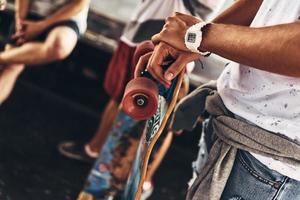 verliebt in Extremsportarten. Nahaufnahme eines jungen modernen Mannes, der sich auf sein Skateboard lehnt, während er Zeit mit Freunden im Freien verbringt foto