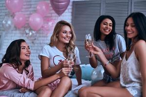 feiern ihre Freundschaft. vier attraktive junge lächelnde frauen in pyjamas, die sich gegenseitig anstoßen, während sie eine schlafparty im schlafzimmer veranstalten foto