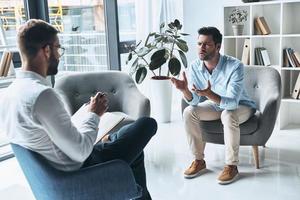 versuchen zu helfen. junger frustrierter mann, der spricht und gestikuliert, während er auf der therapiesitzung mit dem psychologen sitzt foto