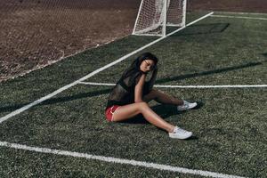 Schönheit auf dem Fußballplatz. Schöne junge Frau in Sportkleidung, die in die Kamera blickt, während sie auf dem Fußballplatz sitzt foto