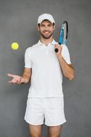 fertig zu spielen. Fröhlicher junger Mann in Sportkleidung, der Tennisschläger auf der Schulter trägt und Tennisball wirft, während er vor grauem Hintergrund steht foto